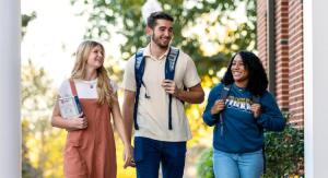 Three college students walking and smiling together on a campus pathway surrounded by greenery and brick buildings.