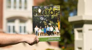 A hand holding the Hilltop Magazine 2025 edition in front of a red brick building and green trees on a sunny day.