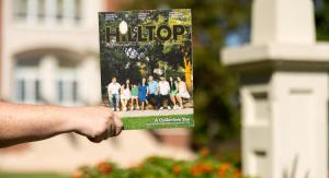 Picture of a hand holding the Hilltop 2025 Magazine in front of a brick building on a sunny day with plants blurred in the background.