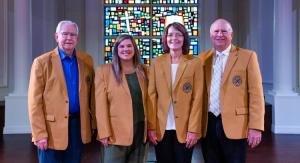 Group of four men and women standing in front of a stained glass window wearing golden blazers.