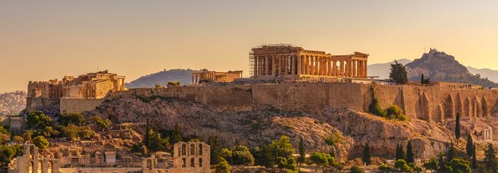 A wide shot of the ancient Acropolis in Athens, Greece, bathed in warm, golden sunlight. The Parthenon and other classical structures dominate the hilltop, with surrounding greenery and the cityscape in the background.