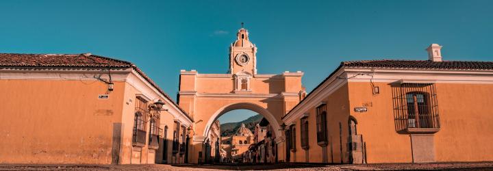 A low-angle view of the Santa Catalina Arch, a historic landmark in Antigua, Guatemala. The yellow-colored archway, topped with a clock, spans a cobblestone street lined with colonial-style buildings. The image is set against a clear blue sky, with the distant view of the Volcán de Agua visible through the arch.
