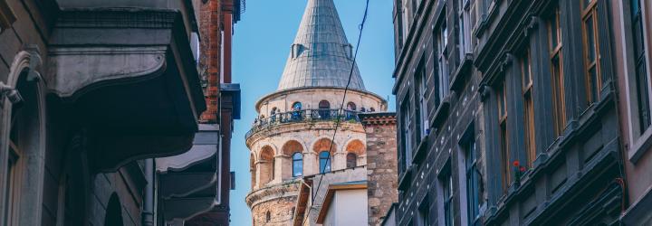 The Galata Tower in Istanbul framed between narrow streets and tall historic buildings, with flower boxes and a clear blue sky overhead.