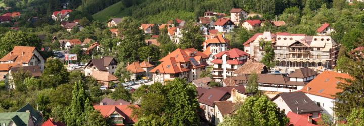 Photograph of a small town nestled in a green, hilly landscape under a blue sky with scattered clouds. The town features numerous houses with red and brown roofs, surrounded by dense trees and vegetation, highlighting a peaceful rural setting.