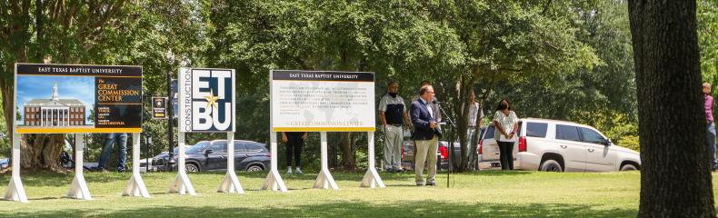 People in a park area with large signs and a white vehicle under trees.