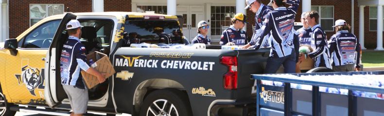 People in uniforms unload a Chevrolet truck and trailer in front of a building with "Evelyn Lineberry Hall" written above the entrance.