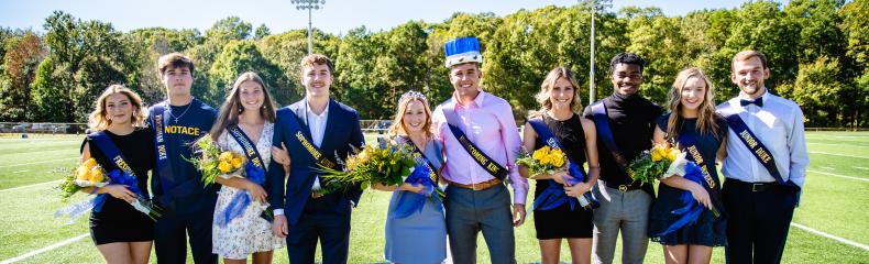 A group of ten people on a sports field, dressed semi-formally, some holding bouquets and wearing sashes.
