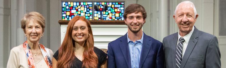Four people standing indoors, two holding awards, with a stained glass window behind them.