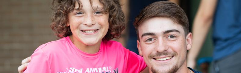 A young person in a pink t-shirt stands next to an adult in a dark shirt, both smiling.
