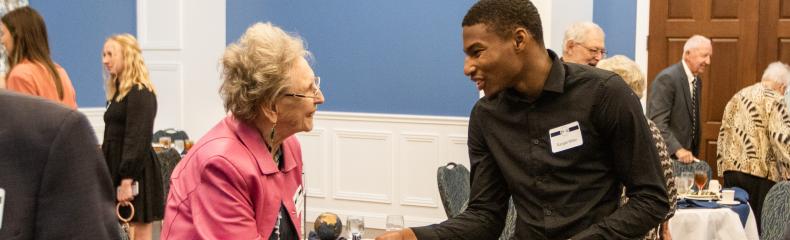 An elderly woman and young man shaking hands at a formal gathering in a blue-walled room.