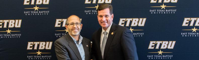 Two men shaking hands in front of an "ETBU" backdrop with documents on the table.