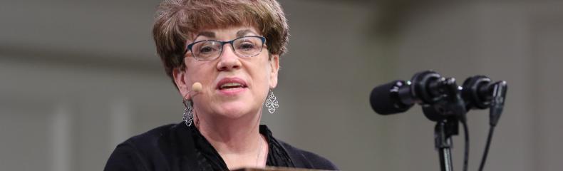 A person with short hair and glasses speaks at a podium with multiple microphones.
