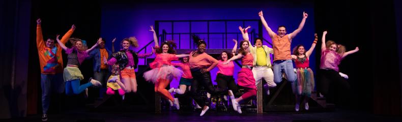 Performers in colorful outfits jumping on a stage with a vibrant purple and blue background.