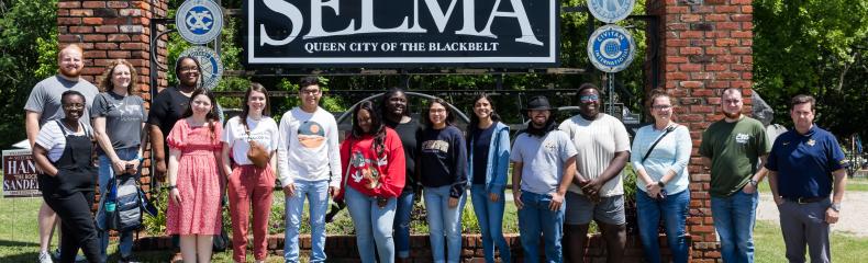 A group of nineteen people stands in front of a sign reading "Welcome to Historic Selma, Gateway to the Blackbelt," with trees in the background.