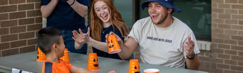 A man and woman sitting at a table with pumpkin cups smiling at a little boy with a woman clapping her hands in the background