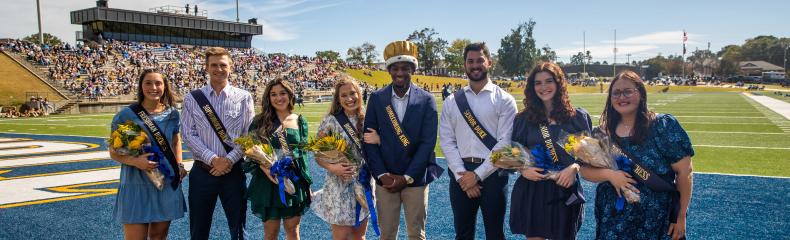 Group of men and women smiling at camera with sashes on and the women holding flowers at the football field