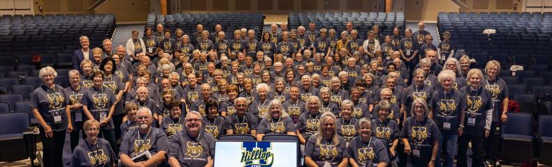 Large group of people in an auditorium wearing matching grey t-shirts, with a screen in front displaying a logo.