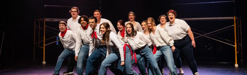 A group of performers in white shirts and blue jeans energetically pose on a stage with colorful lighting.