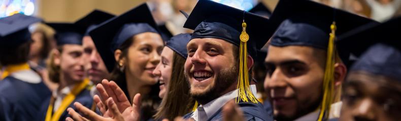 Graduates in navy caps and gowns clapping during a ceremony.