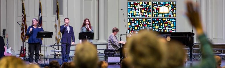 Men and women on stage singing with people worshipping in the foreground in a chapel