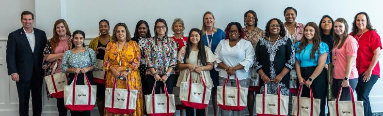 Group of females and one man smiling at the camera holding white and pink tote bags inside