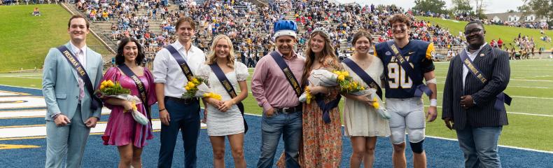  A group of nine people posing on a sports field during an event, some holding bouquets and wearing sashes.
