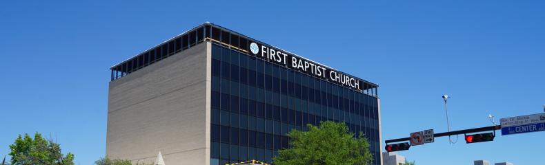 Street corner view of a beige commercial building with dark-tinted windows and "FIRST BAPTIST CHURCH" signage, surrounded by trees and with vehicles on the roads.