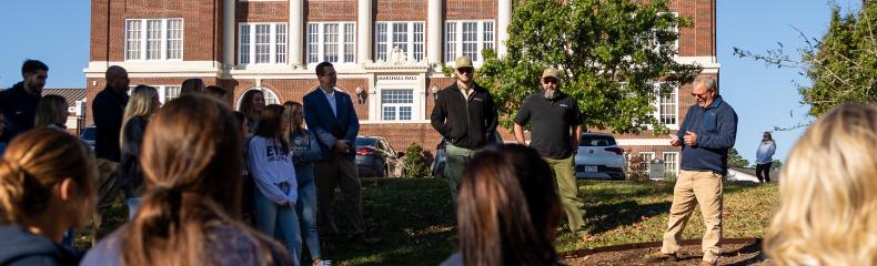 A group of people standing in front of a large brick building, listening to a speaker.