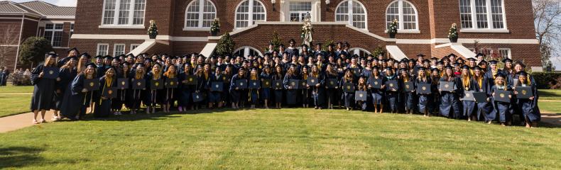 Large group of graduates posed with diplomas in front of brick academic building smiling at the camera