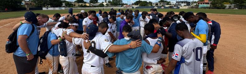 Baseball players praying in a circle with their arms around each other outdoors on a baseball field