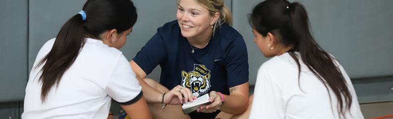 A college student sitting on a gym floor reading with two grade school students