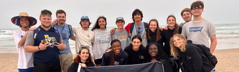 A group of individuals pose on the beach holding a flag with the ETBU logo on it