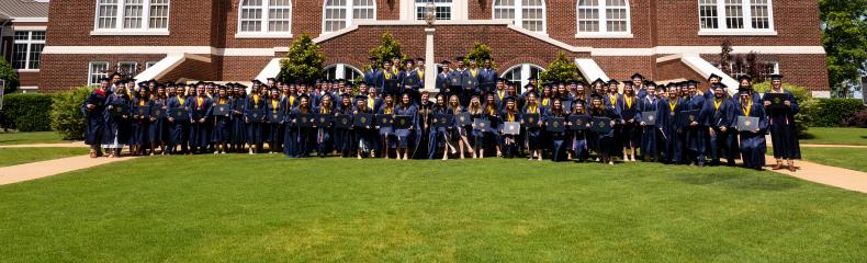 A group of college graduates in navy caps and gowns hold their degrees in front of a red brick building