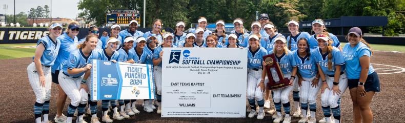 A large group photo of female softball players and coaches with a trophy in front of a score board on Taylor Field at East Texas Baptist University.