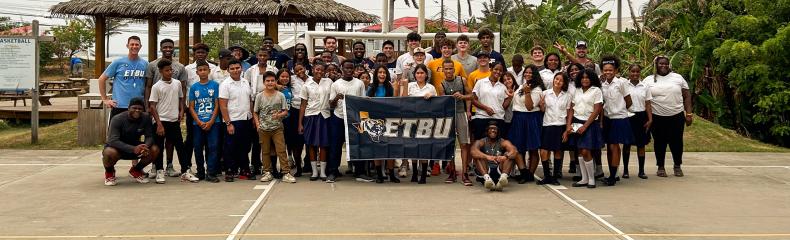 ETBU Men's Basketball Team stand on a basketball court with youth in Honduras holding an ETBU-branded flag