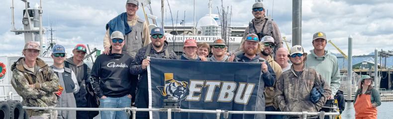 A group of men on a docked fishing boat called the Whistler holding an ETBU navy flag.
