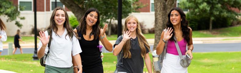 Four young women walking on a college campus, smiling and waving towards the camera. They are dressed in casual attire and carrying backpacks. The background shows green lawns, trees, and part of a brick building.