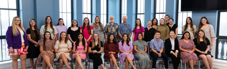 Group photo of a diverse team of individuals at East Texas Baptist University, posed in a bright, spacious room with large arched windows. The group consists of men and women, dressed in a variety of professional and semi-casual attire, seated and standing in two rows.