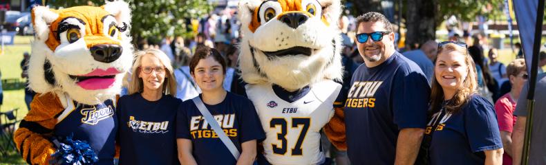 A group of people, including two university mascot tigers, pose together at an outdoor campus event, all wearing school spirit shirts.