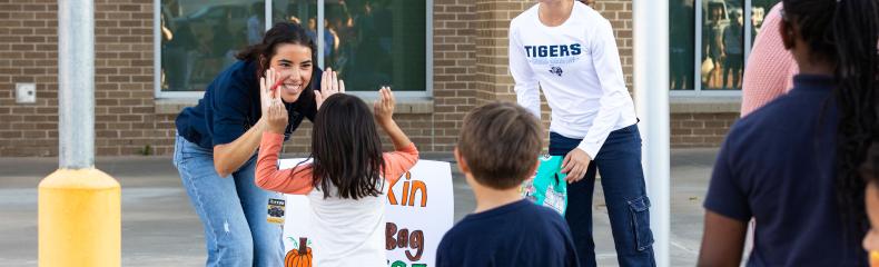Two volunteers engage with children at a fall festival activity, sharing high-fives in front of a sign that reads "Pumpkin Bag Toss."