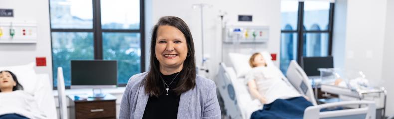 Smiling woman in a professional outfit stands in a modern nursing simulation lab, with hospital beds, mannequins, and medical equipment visible in the background.