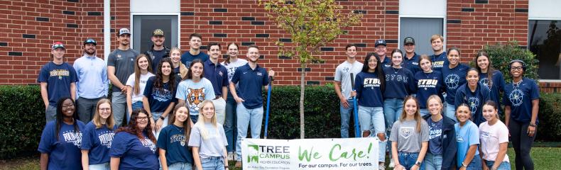 Students stand around a freshly planted tree with a banner between them in honor of Texas Arbor Day