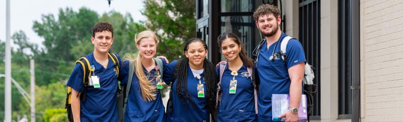 Five students stand side by side outside of building in nursing school scrubs, some holding textbooks and carrying backpacks