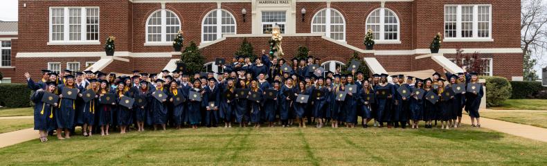 A large group of graduates in blue robes poses in front of a historic red-brick university building.