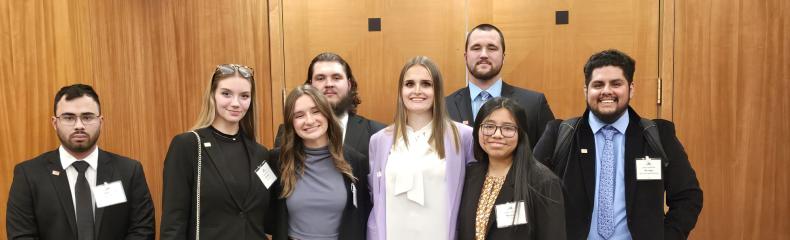 Eight students stand side by side in business professional attire with name badges on.