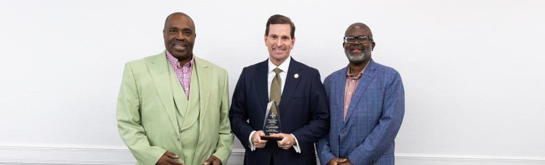 Three men are standing together in a formal setting, with the man in the center holding an award.