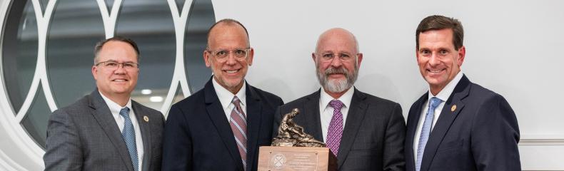 Four men in suits stand together, one holding a statue with a wooden base.