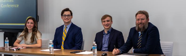 Four people sit behind a table in professional attire, a TV screen with a presentation is seen in the background
