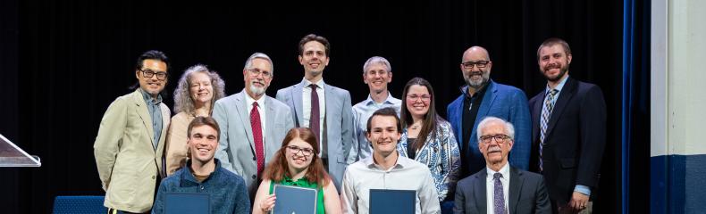 A group of people sitting and standing on a dimly lit stage smiling for a group photo