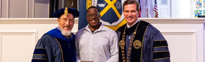 3 people stood in front of the seal for East Texas Baptist University, in a well-lit room wearing academic regalia
